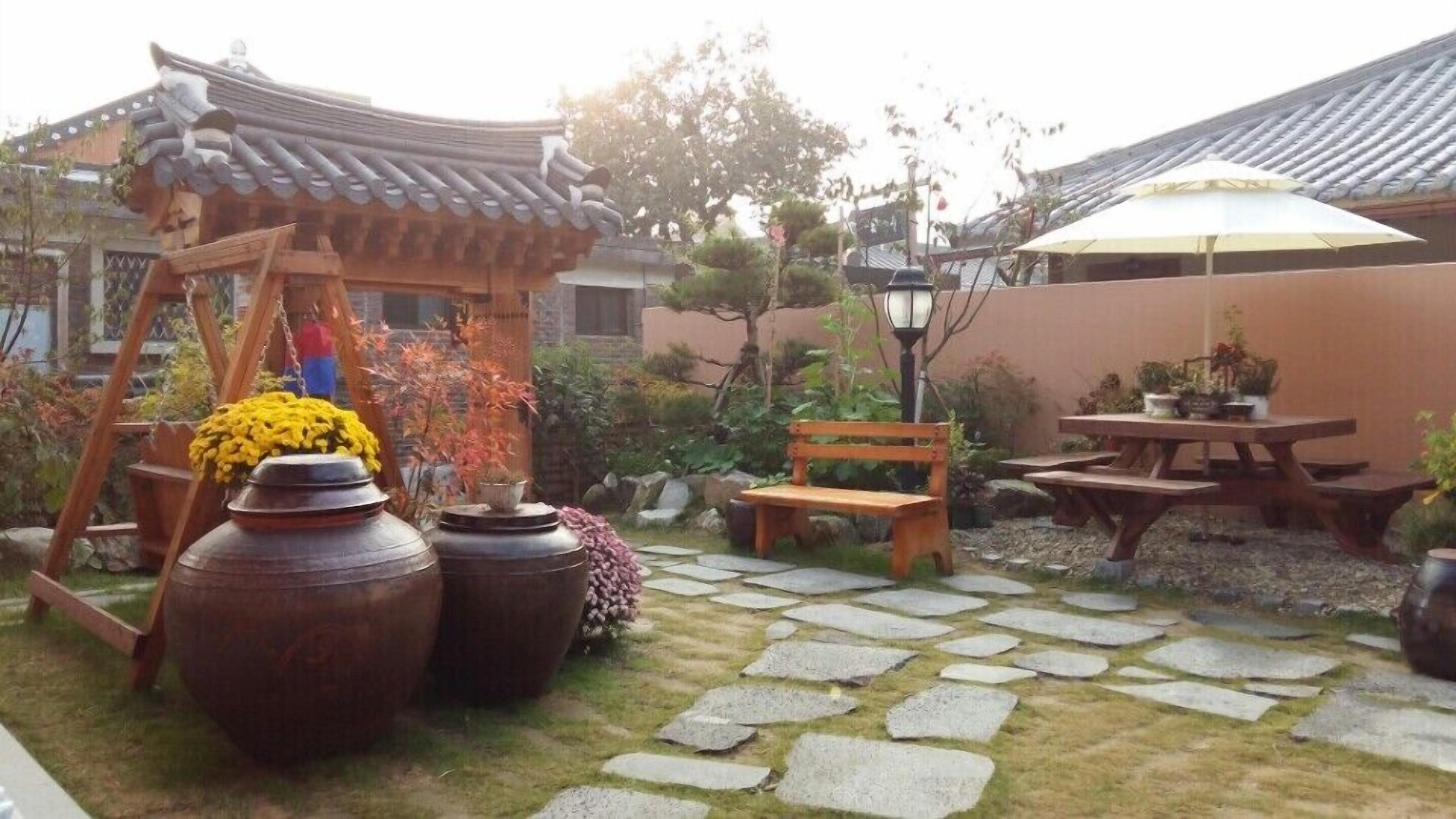 Un jardin traditionnel de style coréen, avec des allées pavées de pierres, un pavillon en bois, un banc et une table agrémentée de plantes en pot.