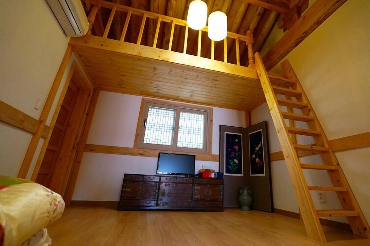 A loft bedroom with a wooden ceiling, a window, a dresser, and a ladder.