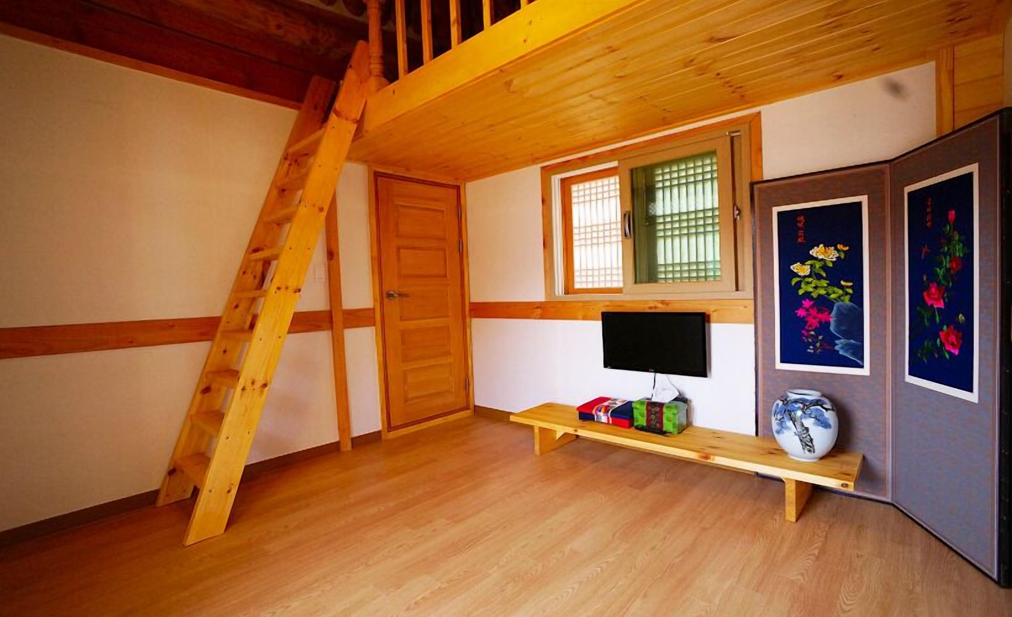 A room with wooden beams, a wooden ladder, a TV, a shelf with items, and a window with blinds.