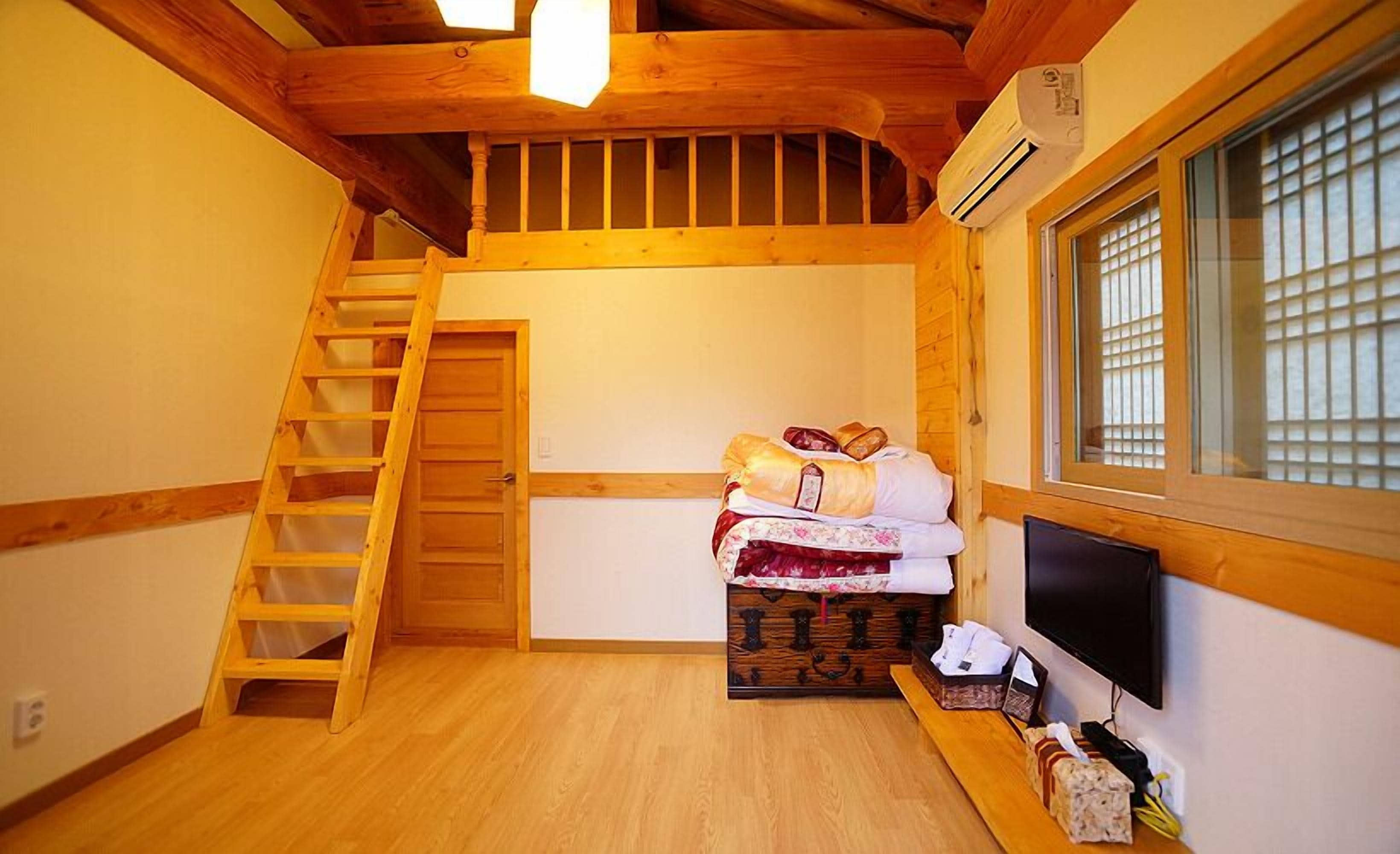 A small, cozy room with a wooden ladder leading to a loft, a bed with pillows and blankets, a TV, and a window with blinds.