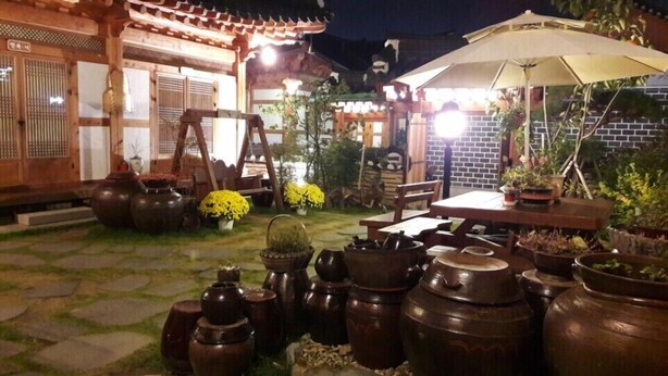 An outdoor patio with traditional Korean architecture, including a wooden building with tiled roof, a table with chairs, and various potted plants and traditional Korean pottery. - The Mumum (Jeonju)
