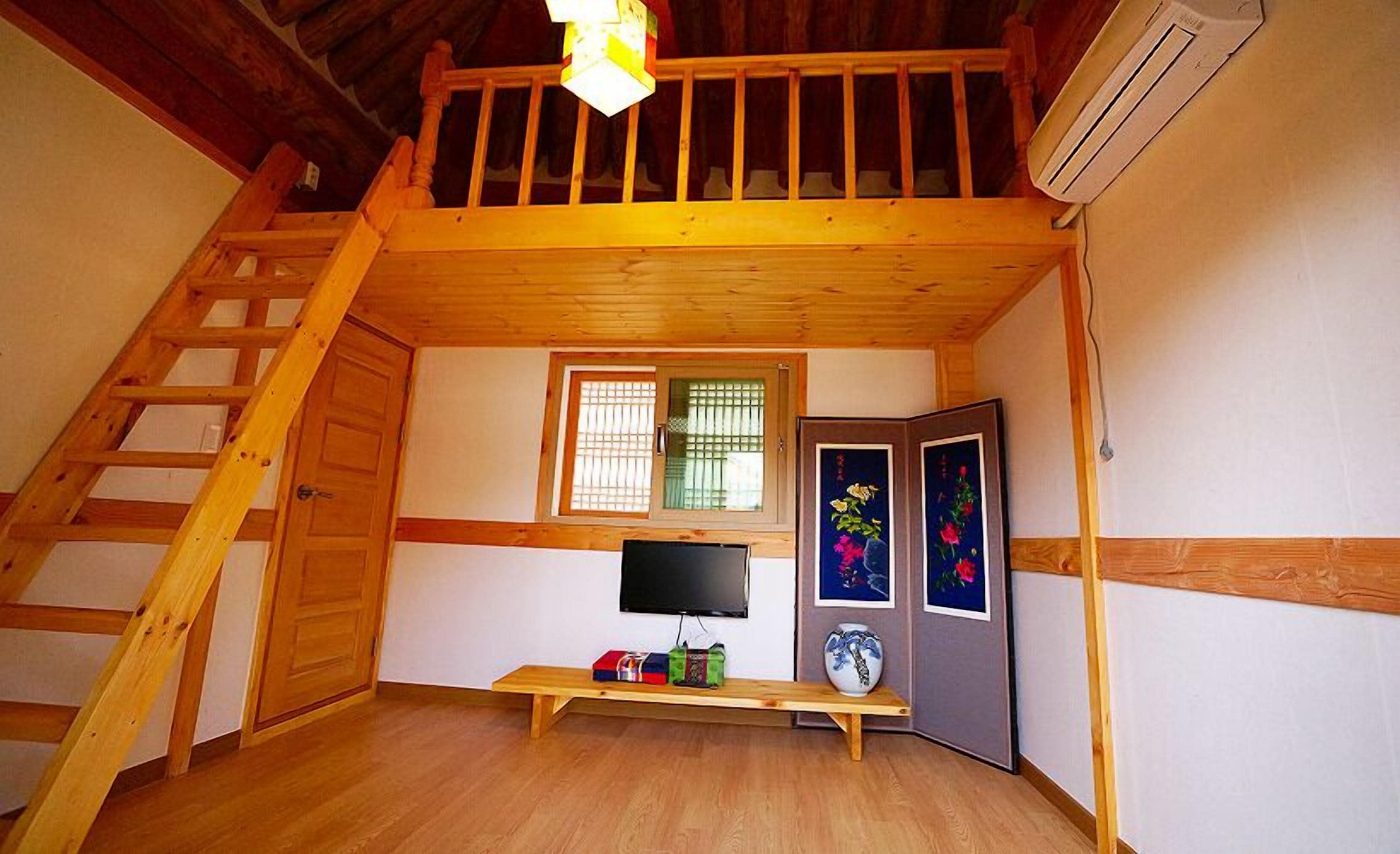 A loft bedroom with a wooden ladder, a TV, a bench, and a window with blinds.