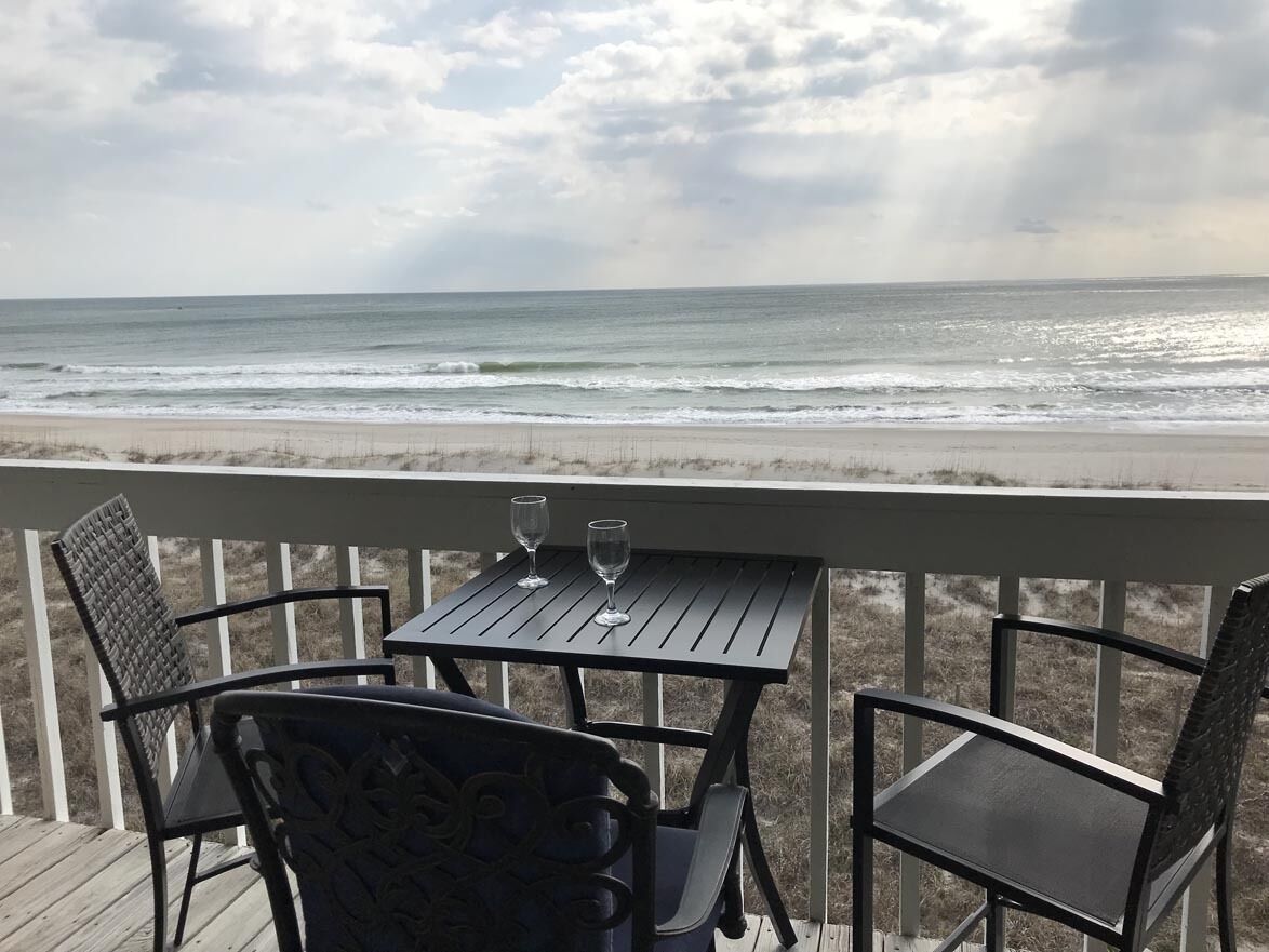 A wooden deck with two chairs and a table, overlooking a beach with waves and a cloudy sky.