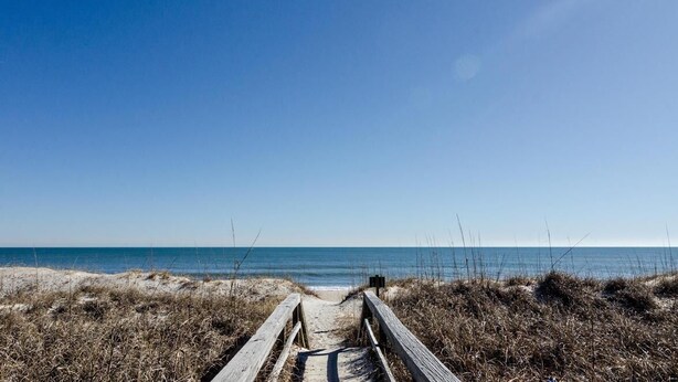 Ein hölzerner Steg führt zu einem Strand bei blauem Himmel.