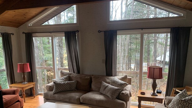 A cozy living room with a sofa, a coffee table, and a lamp. There are large windows with curtains, and a view of trees outside. - Great Vacation Destination, 1 Mile To Storyland, Skiing, Golf, Hiking. (Bartlett)
