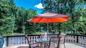A wooden deck with a glass table and chairs, a red umbrella, and a view of a forested area. - Great Vacation Destination, 1 Mile To Storyland, Skiing, Golf, Hiking. (Bartlett)