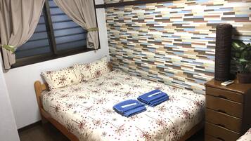Un dormitorio con una colcha estampada, un cabecero de madera, una ventana con cortinas, una pared con un diseño pixelado, un aparador con un control remoto y una planta.