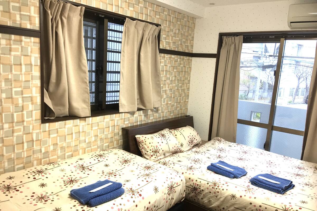 A hotel room with two beds, a window with curtains, a tiled wall, and a view of the street.