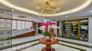 A lobby with a marble floor, a round wooden table with a vase of flowers, a chandelier, and a reception desk with a patterned wall. - Manli Resort Quang Binh (Dong Thuan)