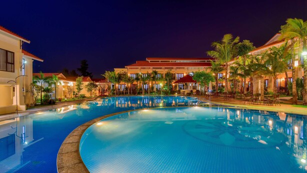 A nighttime swimming pool area with a curved edge, surrounded by palm trees and buildings with warm lighting. - Manli Resort Quang Binh (Dong Thuan)