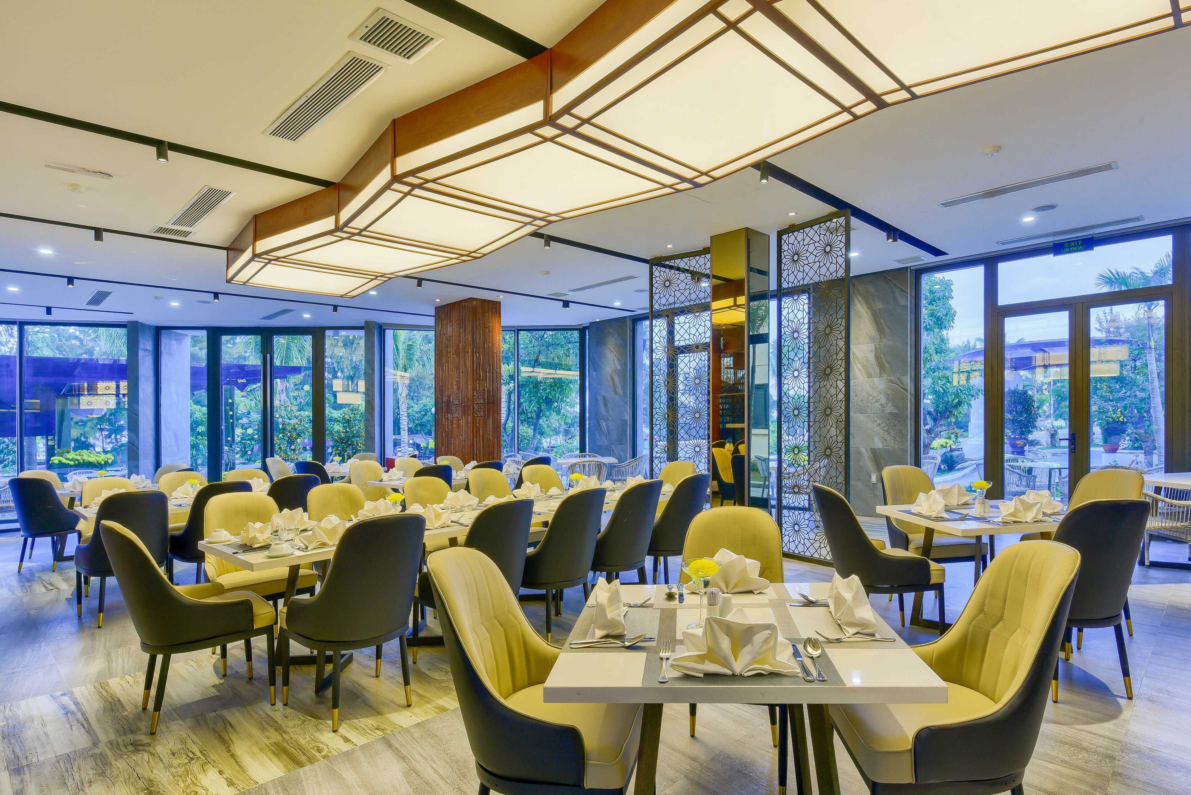 A spacious dining area with multiple tables and chairs, large windows offering a view of the outdoors, and a modern ceiling design with exposed beams and lighting fixtures. - Manli Resort Quang Binh (Dong Thuan)