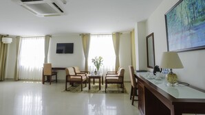 A hotel room with a dining area, a flat-screen TV mounted on the wall, a desk with a lamp, and a painting above the desk. - Luxe Hotel (Dong Hoi)