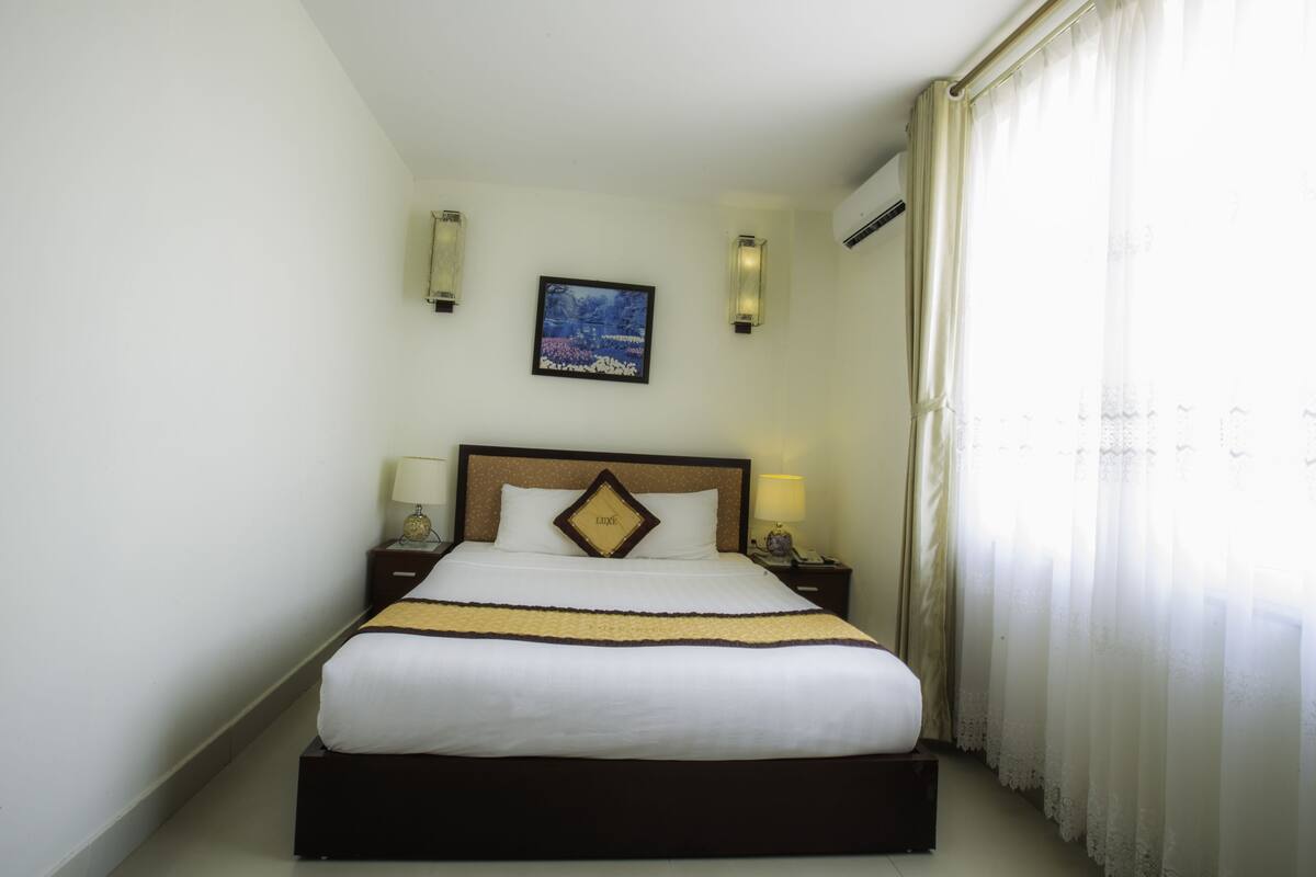 Habitación de hotel con cama, dos lámparas de noche, mesita de noche, un cuadro enmarcado en la pared y una ventana con cortinas.