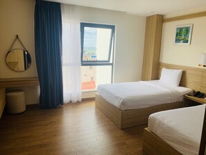 A hotel room with two beds, a wooden floor, a window with a view, a bedside table with a phone, and a mirror on the wall.