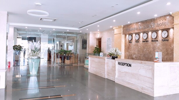 A modern hotel reception area with a marble reception desk, a glass wall leading to a lounge, and clocks displaying different cities. - Luxe Hotel (Dong Hoi)
