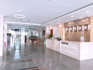 A modern hotel reception area with a marble reception desk, a glass wall leading to a lounge, and clocks displaying different cities. - Luxe Hotel (Dong Hoi)