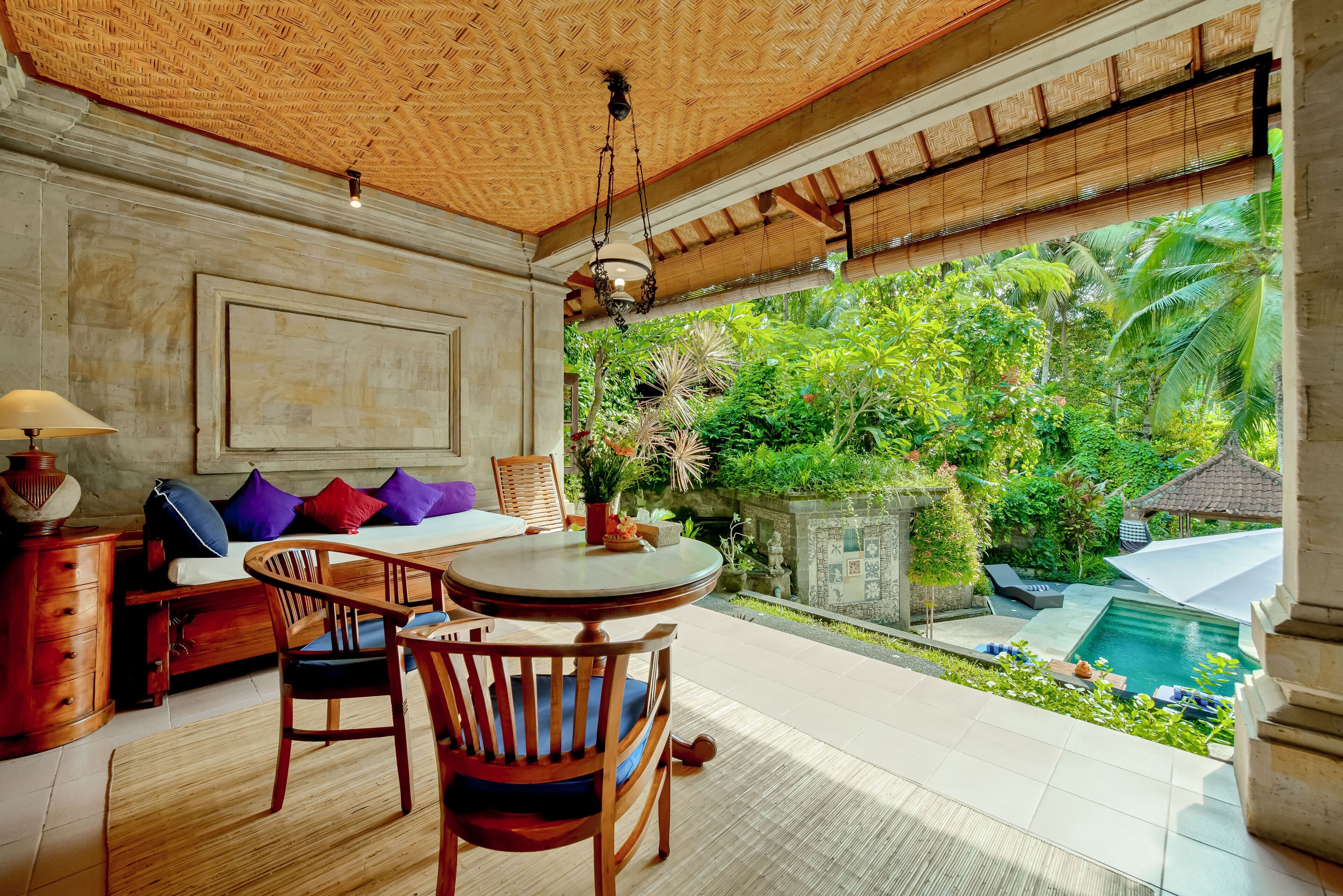A room with a wooden ceiling, a round table with chairs, a sofa with cushions, and a view of a garden and pool outside.