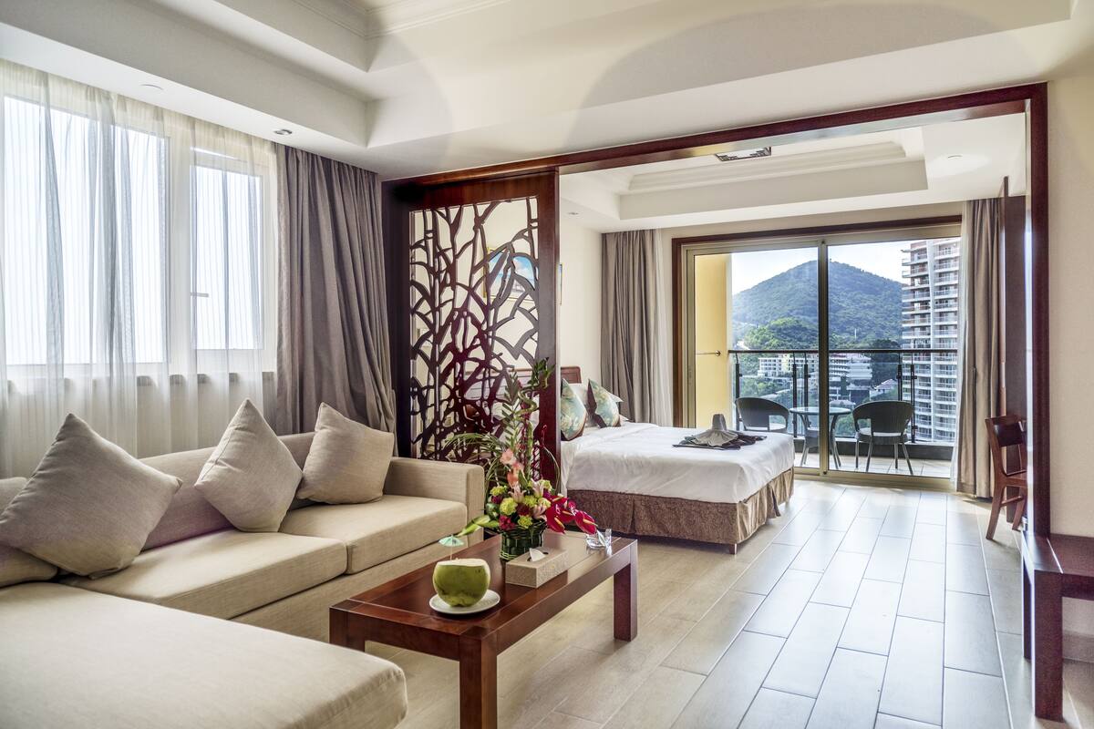 Ein Hotelzimmer mit einer Couch, einem Bett, einem Balkon mit Blick auf Gebäude und Berge sowie einer dekorativen Raumtrennung.