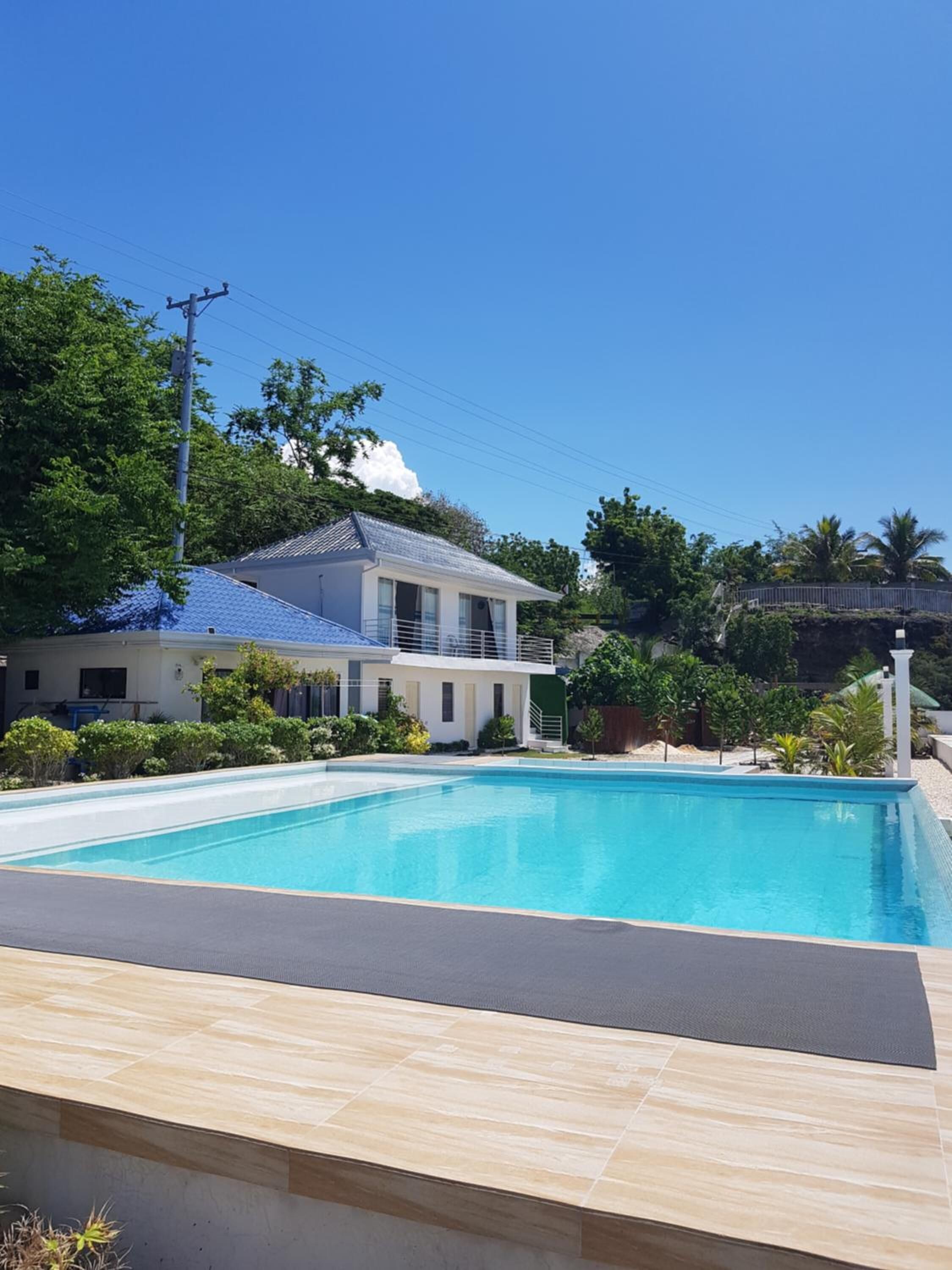 Una piscina con borde de baldosas, una casa con techo azul y follaje circundante.