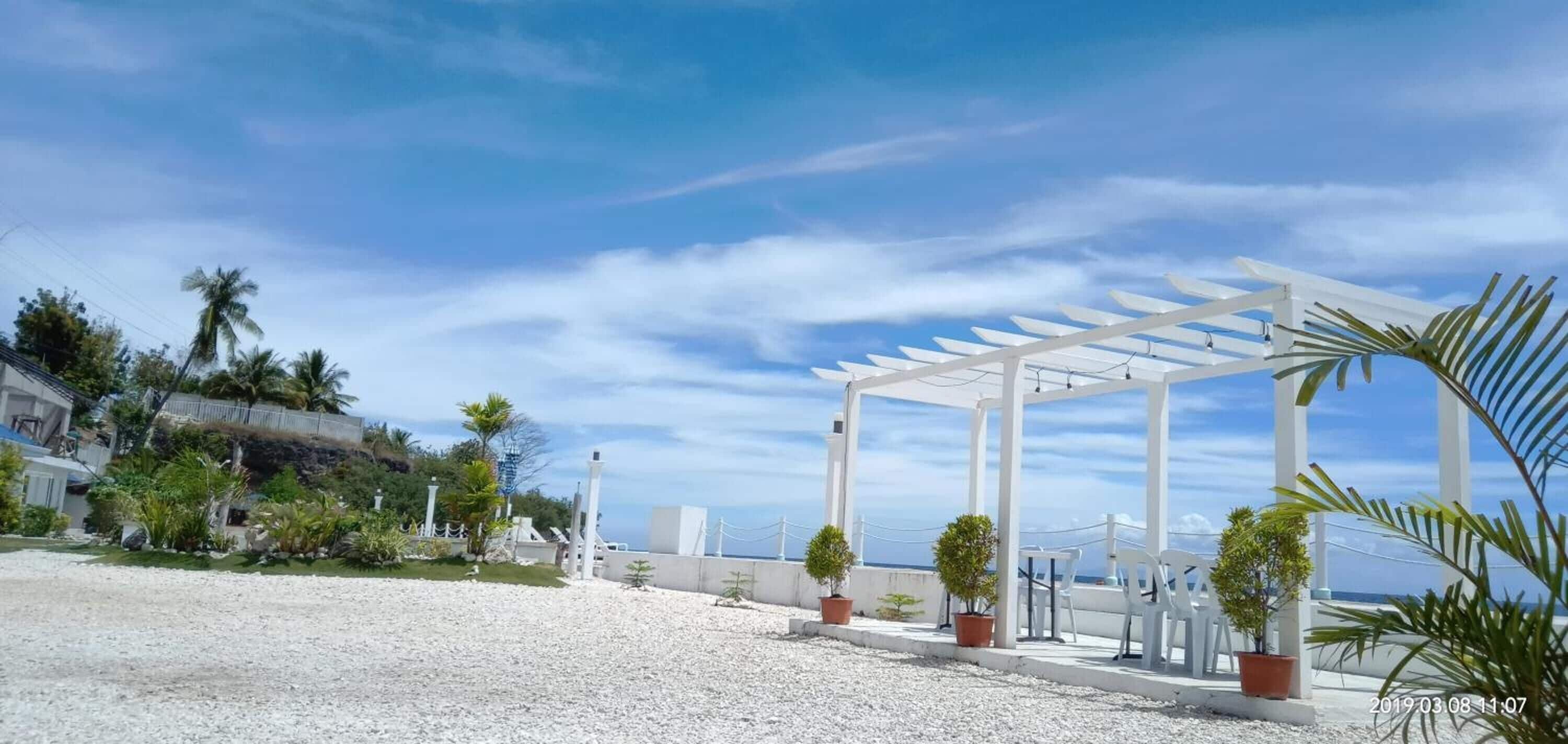 Una estructura de pérgola blanca con plantas en macetas, un área de grava y vistas al mar.
