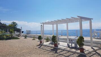 A white pergola with a view of the sea, surrounded by potted plants and outdoor seating.