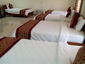 Beds with white mattresses and matching duvets, adorned with a red and gold patterned bedspread, are arranged in a row.
