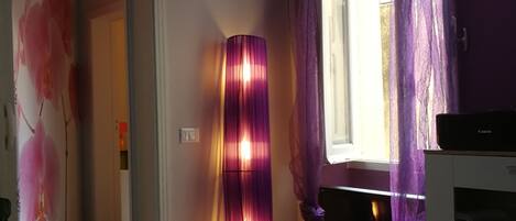 A room with a wooden floor, a standing lamp, a window with purple curtains, and a white air conditioning unit.