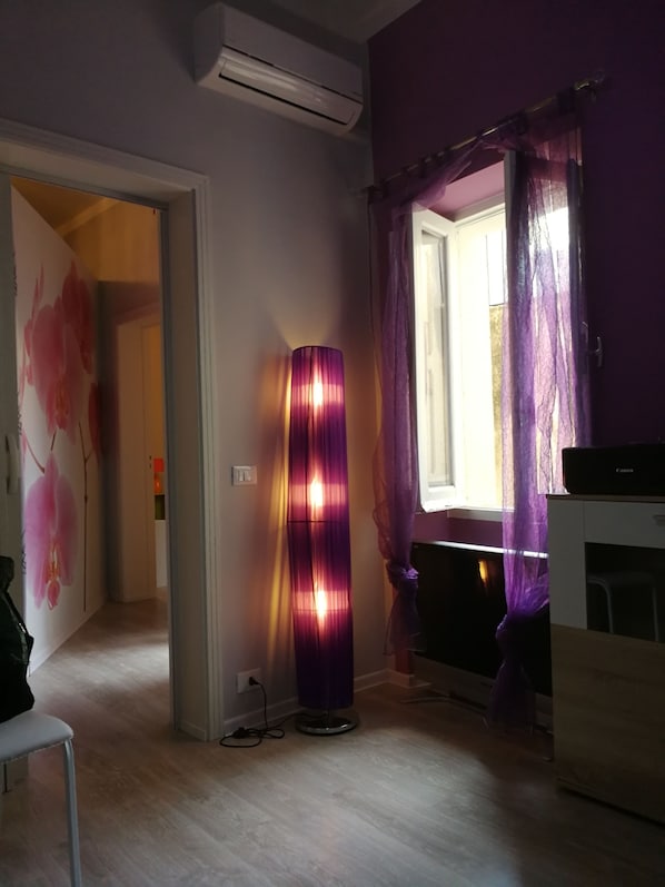 A room with a wooden floor, a standing lamp, a window with purple curtains, and a white air conditioning unit. - BADIA GRANDE APARTMENT (Trapani)