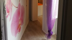 Ein Flur mit floralem Wandbild, einer Tür zu einem Raum mit einem orangefarbenen Tisch und violetten Vorhängen.