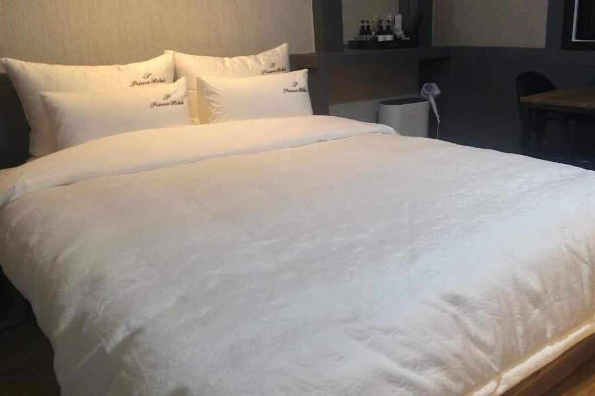 Quarto de hotel com cama grande, roupa de cama branca, piso de madeira, uma mesa pequena e uma cadeira.