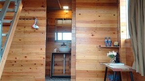 Interior de una cabaña de madera con habitación en el altillo, un baño con lavabo y una pequeña mesa con un florero.