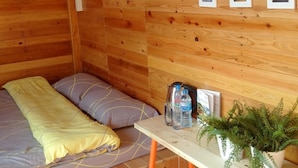 Habitación de cabaña de madera con una cama, una mesita con una botella de agua y una planta, y tres cuadros en la pared.