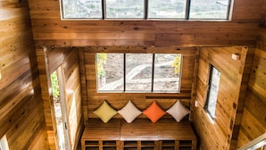 Una cabaña de madera con un dormitorio en el altillo, una pequeña zona de estar con cojines y grandes ventanales con vistas al exterior.
