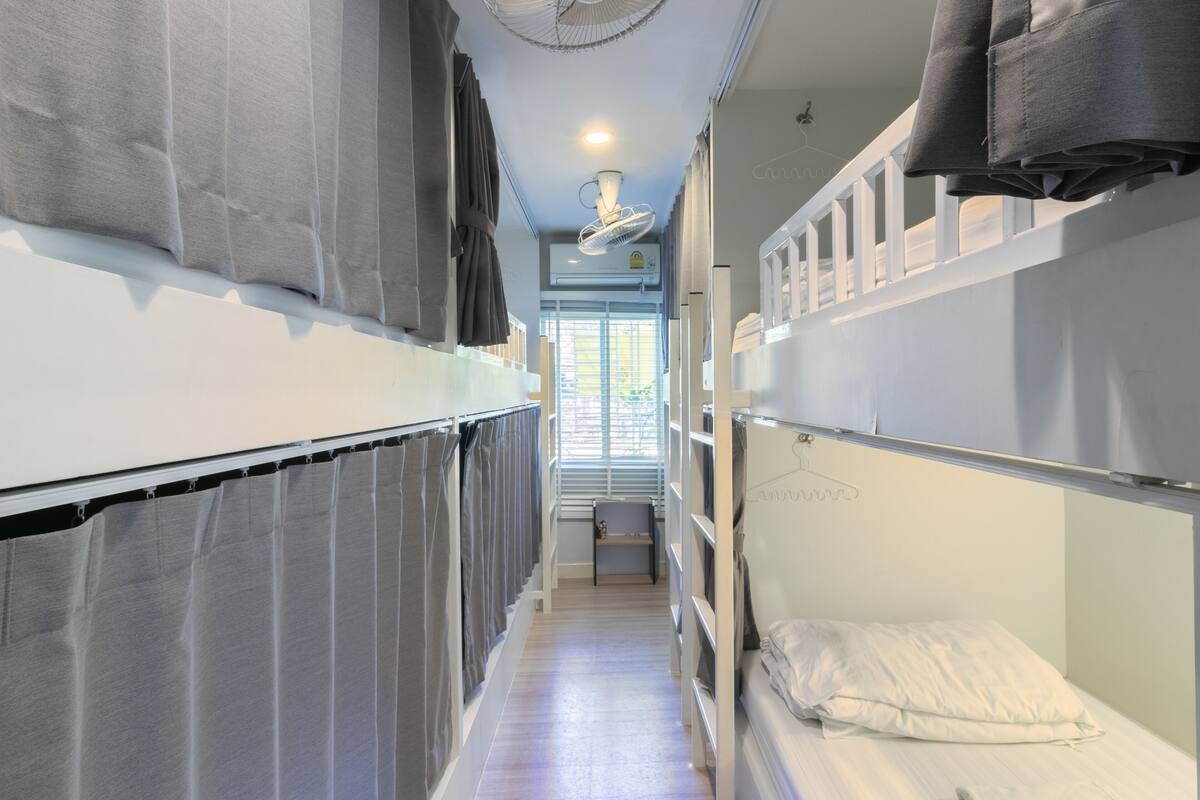 Un couloir étroit, avec des lits superposés de chaque côté, un ventilateur de plafond et une fenêtre avec des stores.