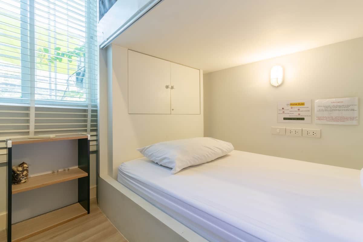 Une petite pièce, bien rangée, avec un lit simple, une table de chevet, une armoire et une fenêtre avec des stores.