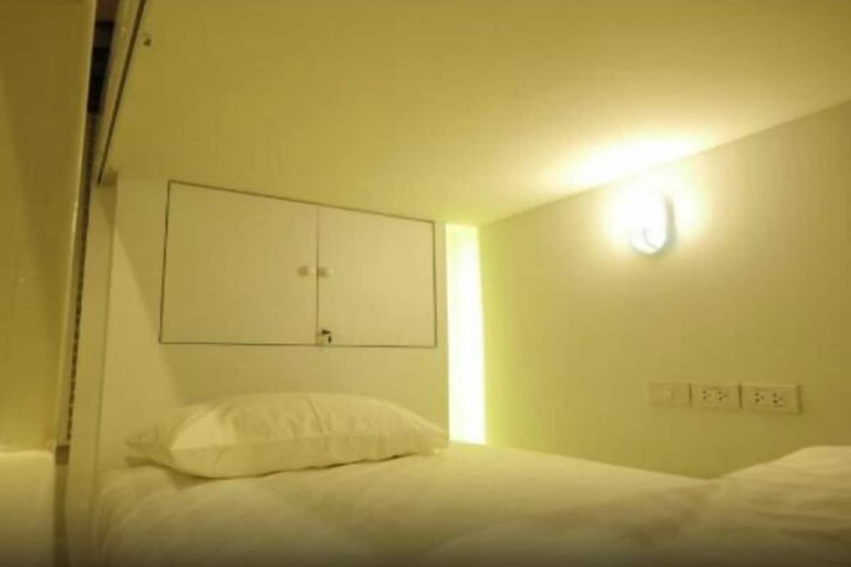 Une petite chambre avec un lit simple, disposant d’un aménagement simple, comprenant un lit, une lampe fixée au mur et une prise de courant.