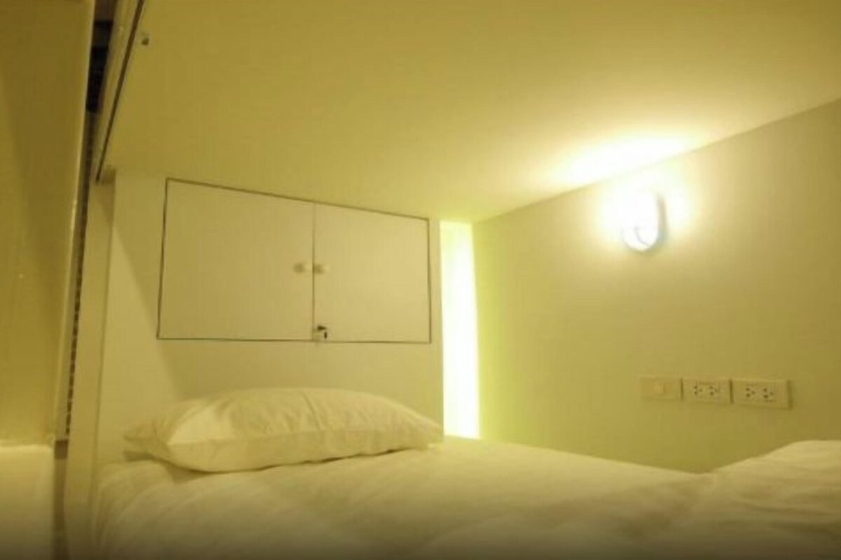 A small, single-bed room with a simple layout, including a bed, a wall-mounted light, and a power outlet.