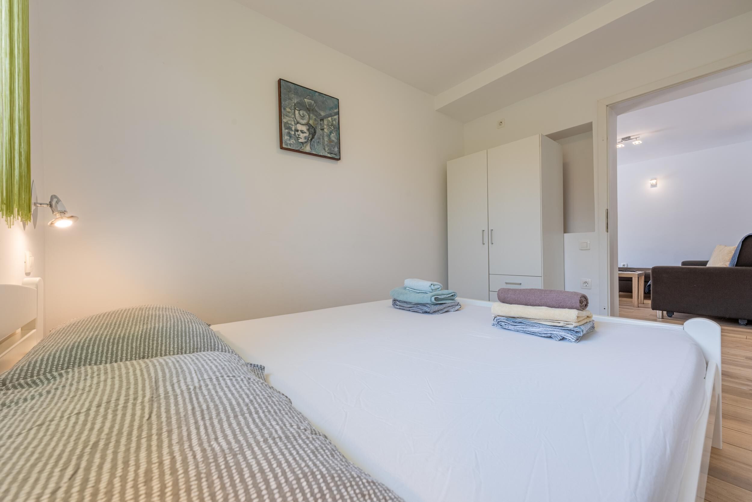 Un lit bien fait, avec des oreillers, une serviette pliée et une couverture sur un matelas blanc. Il y a un tableau au mur et une armoire à l’arrière-plan.