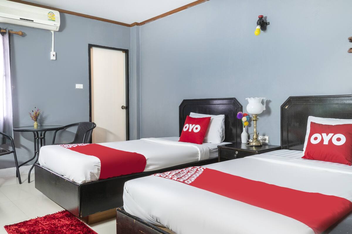 Une chambre d’hôtel avec deux lits, chacun recouvert d’une courtepointe rouge et blanche et agrémenté d’oreillers portant l’inscription « OYO ». On y trouve une petite table avec une lampe et un vase de fleurs, ainsi qu’une chaise.