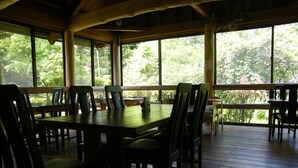 Eine Holzterrasse im Freien mit Esstisch und Stühlen, umgeben von großen Fenstern mit Blick auf Bäume.