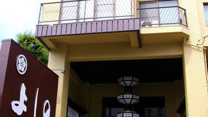 A multi-story building with a prominent entrance, a sign with a logo and text, and a staircase leading to the entrance. - Hotel Yoshino (Ito)