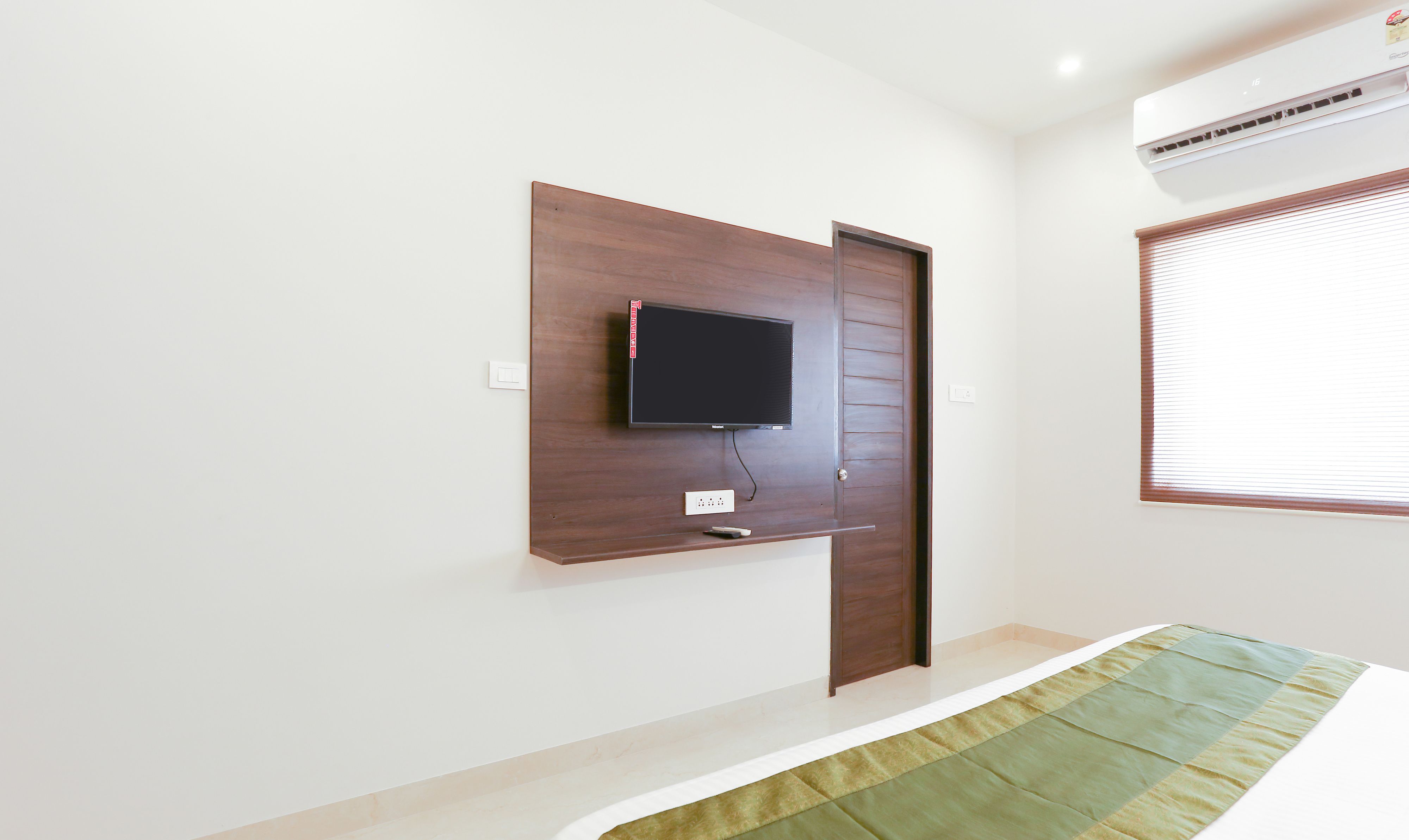 A bedroom with a mounted TV, a bedside table, a power outlet, and a window with blinds.