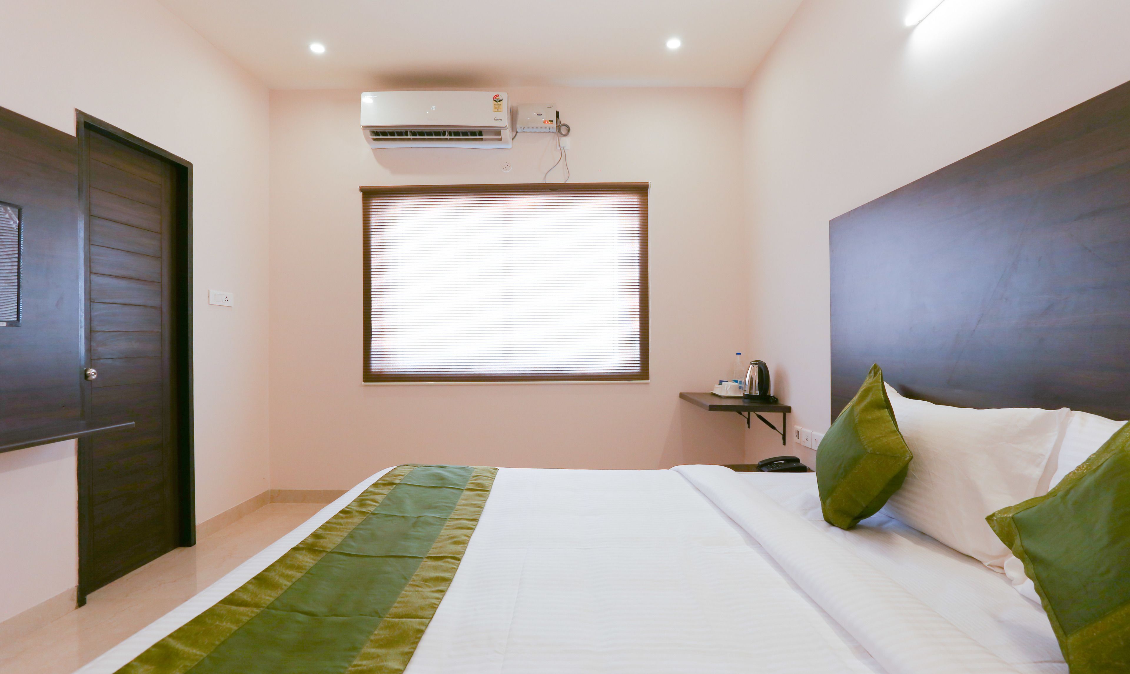 A bedroom with a bed, a window with blinds, an air conditioning unit, a bedside table with a kettle, and a nightstand.