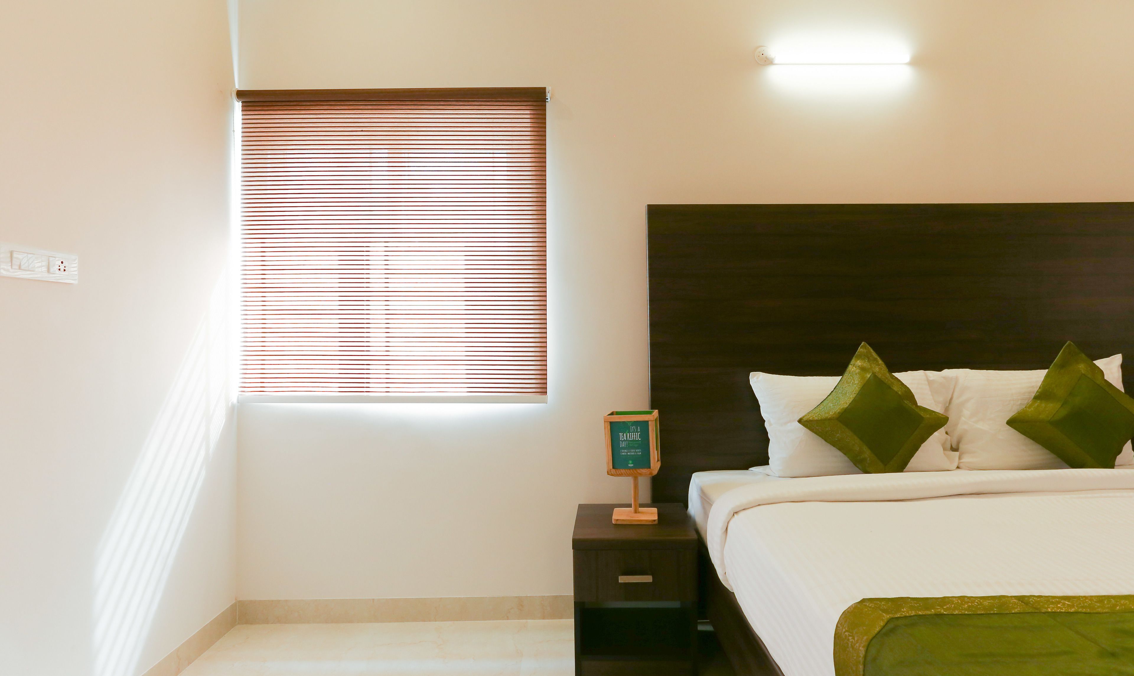 A bedroom with a bed, a nightstand, a window with blinds, and a wall-mounted light.