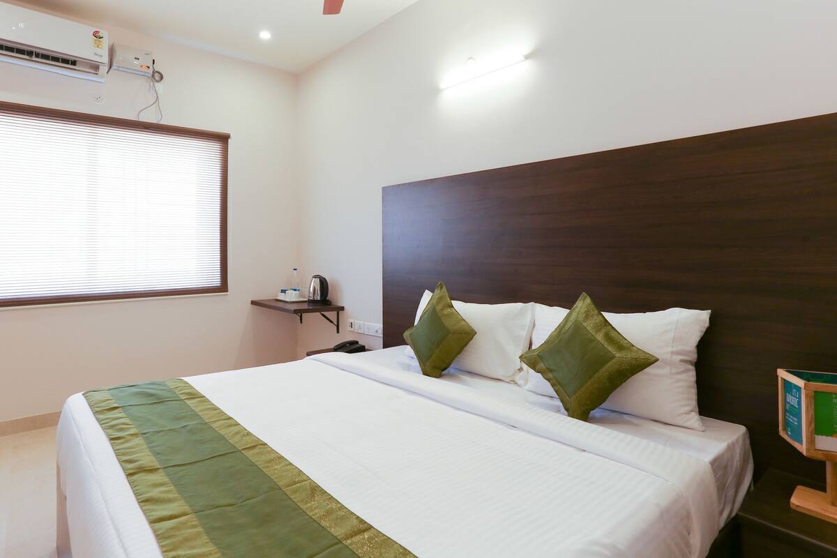 A hotel room with a bed, a nightstand, a window with blinds, and a wall-mounted air conditioner.