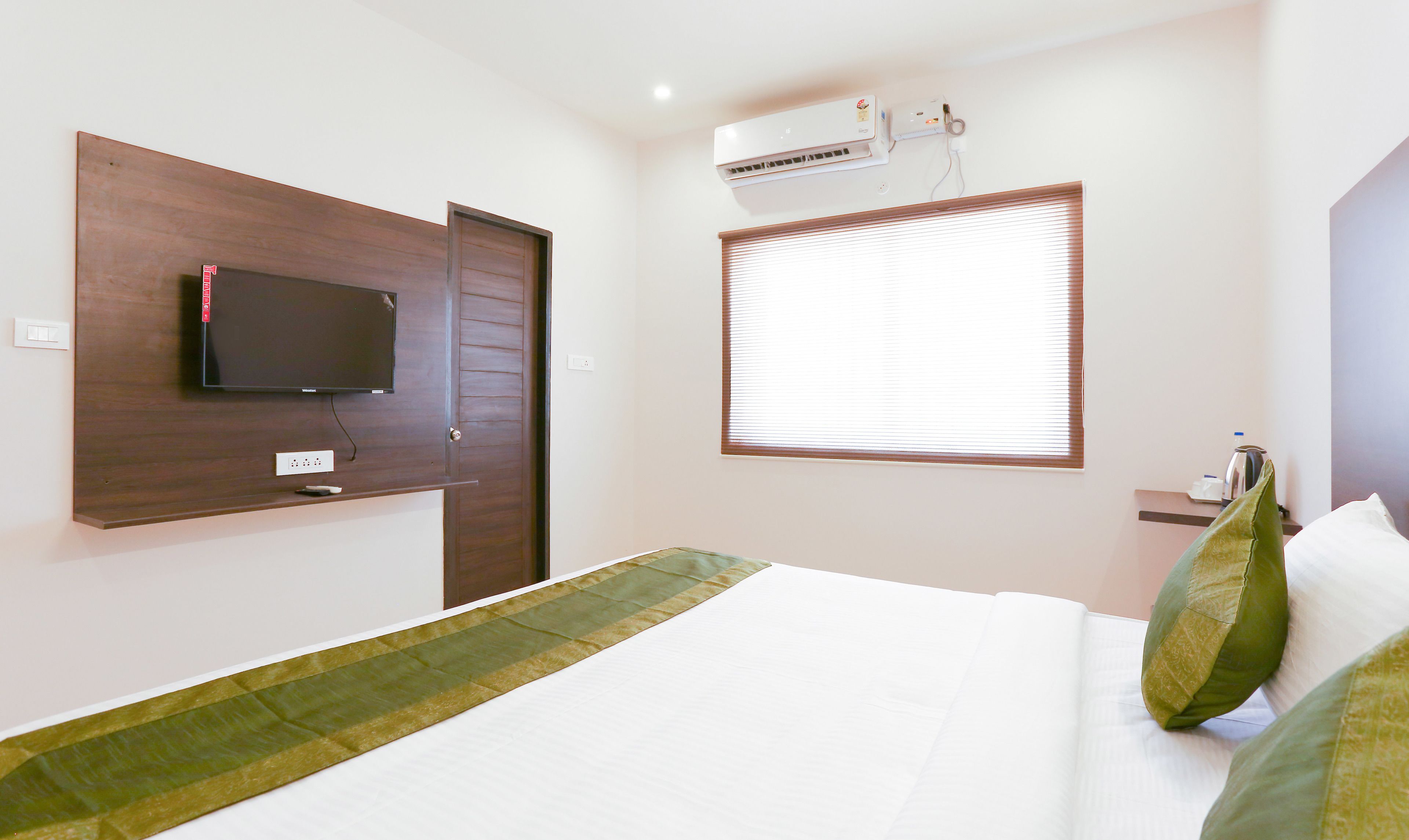 A hotel room with a bed, a television mounted on the wall, an air conditioning unit, a window with blinds, and a small table with a kettle.