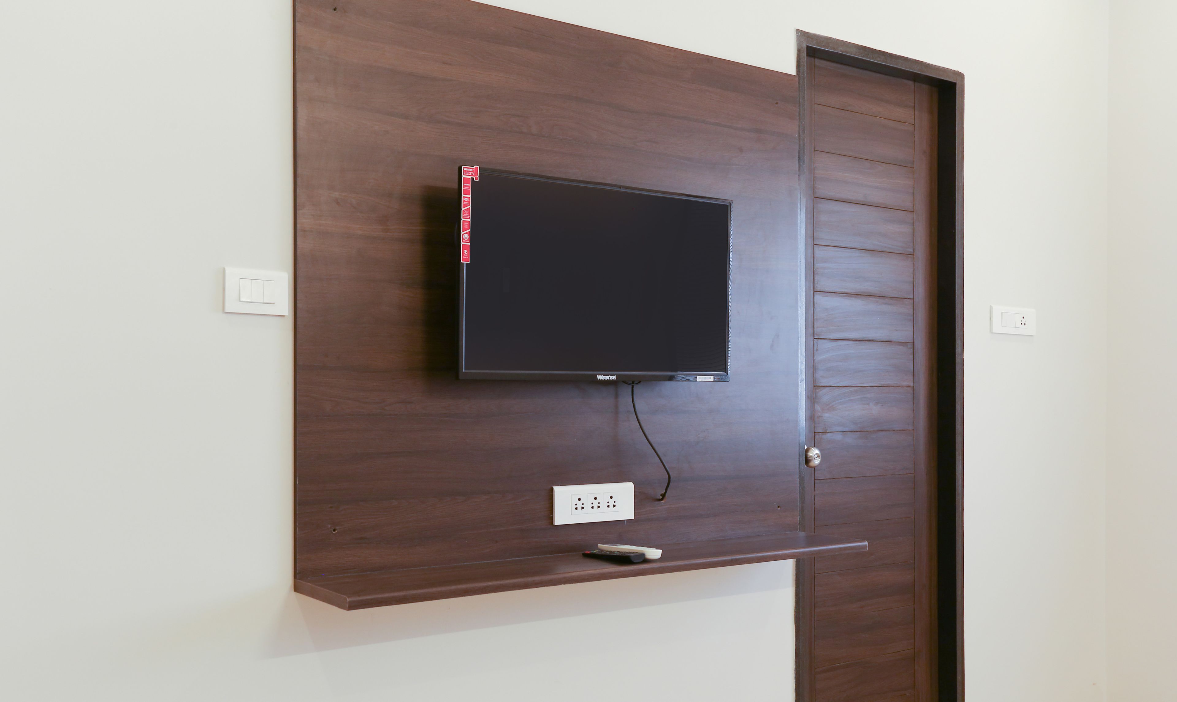 A flat-screen TV mounted on a wooden wall with a power outlet and a remote control below it.