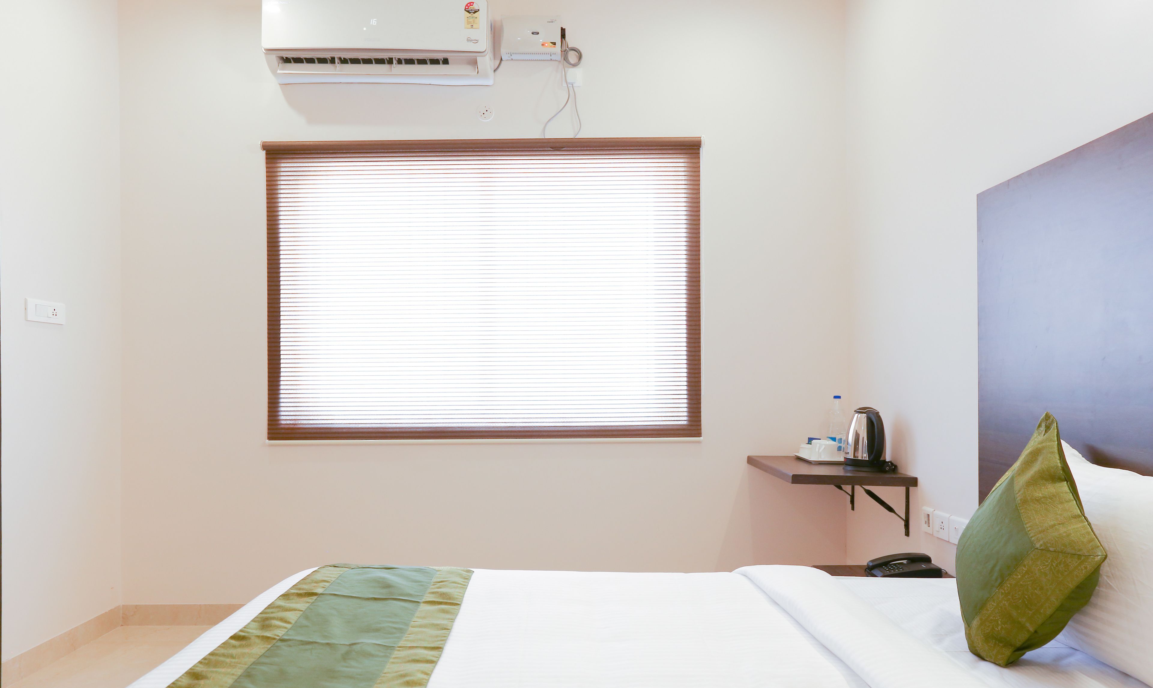 A hotel room with a bed, a bedside table with a kettle, a telephone, and a window with blinds.