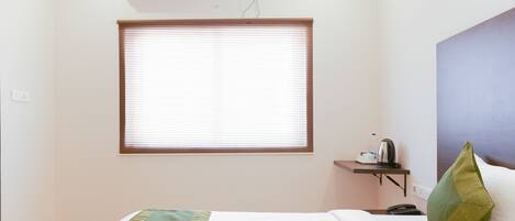 A hotel room with a bed, a bedside table with a kettle, a telephone, and a window with blinds.