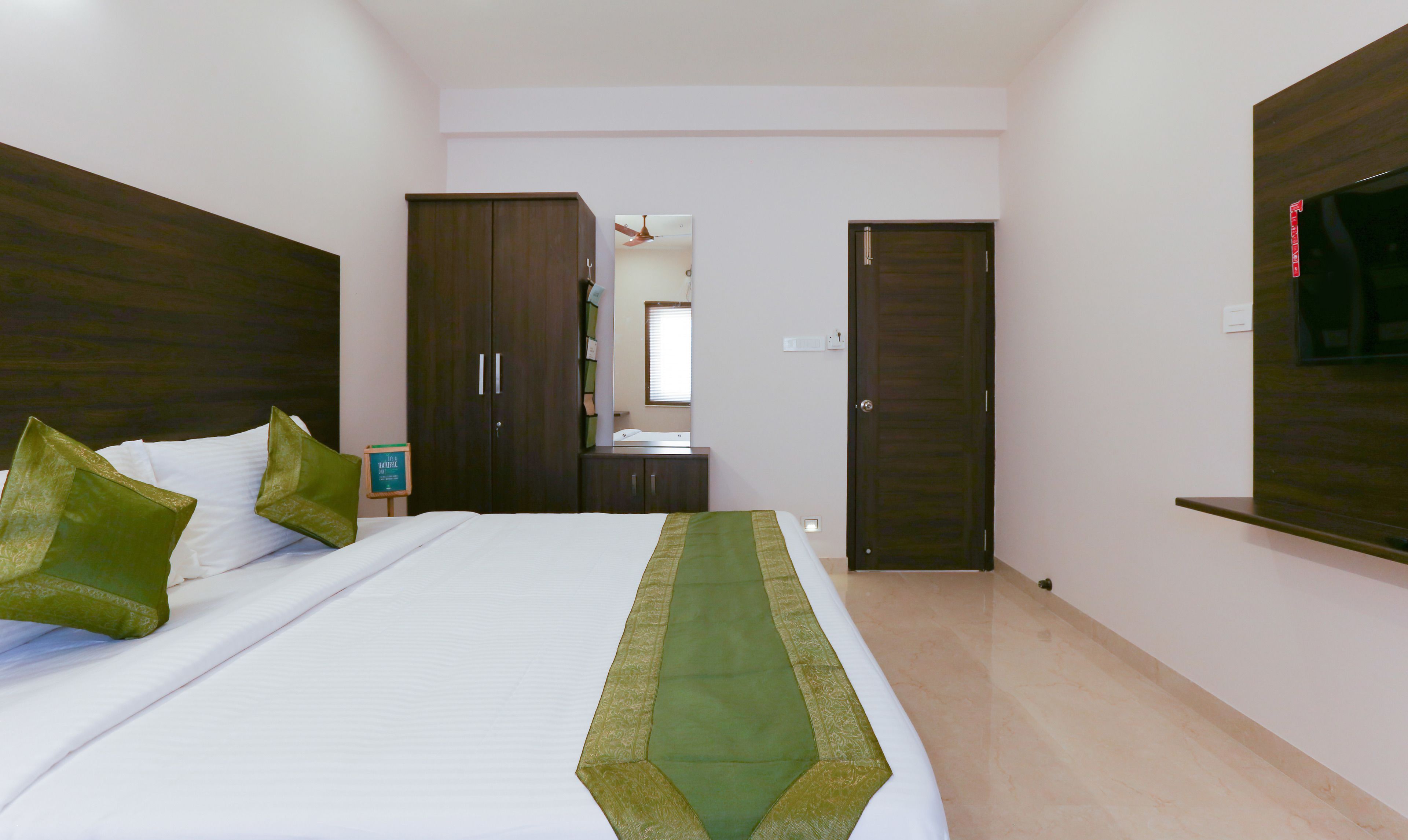 A hotel room with a bed, two green pillows, a nightstand, a mirror, a wardrobe, and a TV mounted on the wall.